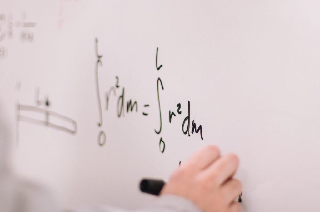 Imagem da mão direita de uma pessoa escrevendo em um quadro operações matemáticas, exercitando suas inteligências múltiplas.