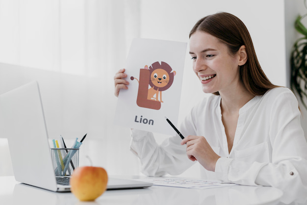 Educadora explica uma das atividades de coordenação motora ampla para seus alunos: em frente ao notebook segura uma folha de papel onde há um desenheo de um leão, a letra L e a palavra "Lion".