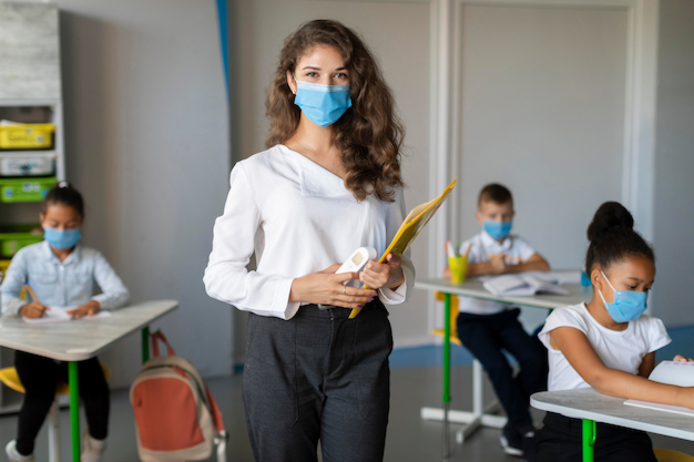 Professora de máscara de proteção realiza acolhimento escolar com os estudantes também de máscara sentados na sala de aula.