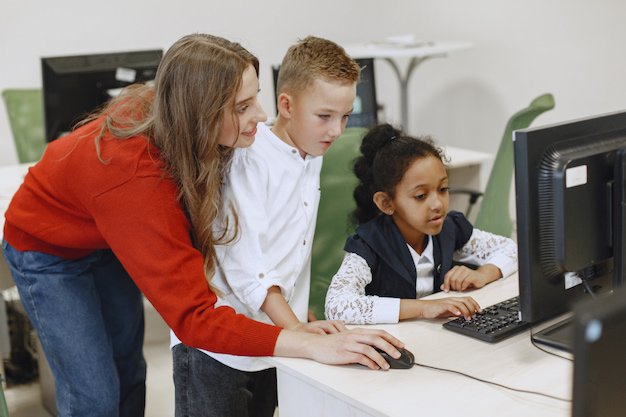 Mulher trabalha o letramento digital na escola com duas crianças em frente ao computador.