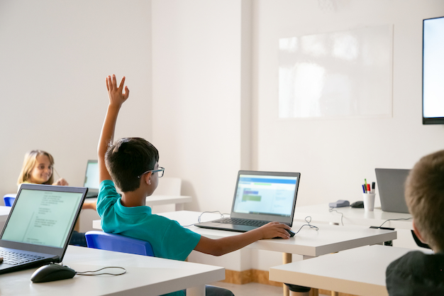 Três crianças em sala de aula em frente aos seus computadores exercitam seu letramento digital na escola. Uma das crianças levanta mão, pedindo para falar.