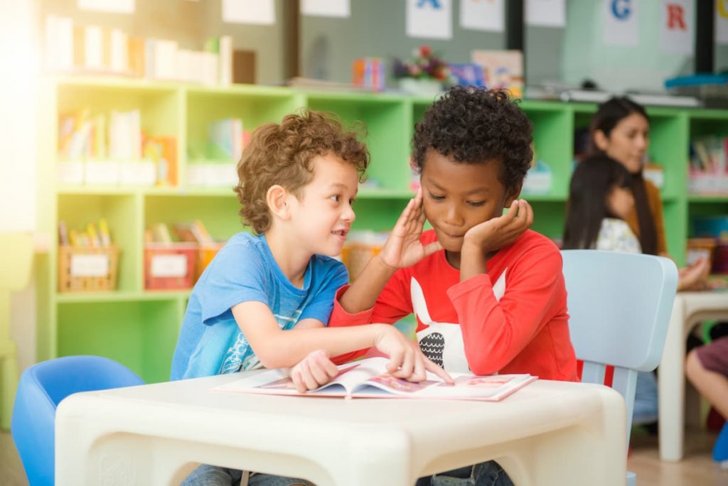 Duas crianças eDuas crianças sentadas em cadeirinhas na sala de aula leem e conversam, ilustrando um momento do ensino híbrido na Educação Infantil.