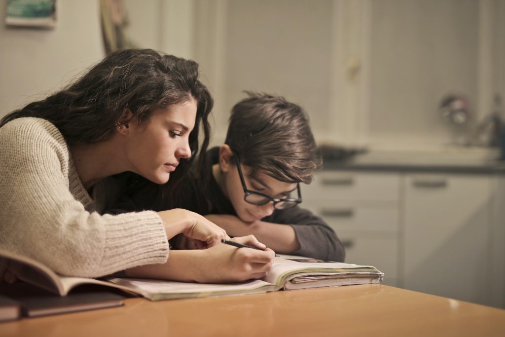 Mulher ajudando uma criança a preencher a lição no livro, demonstrando a relação família e escola.