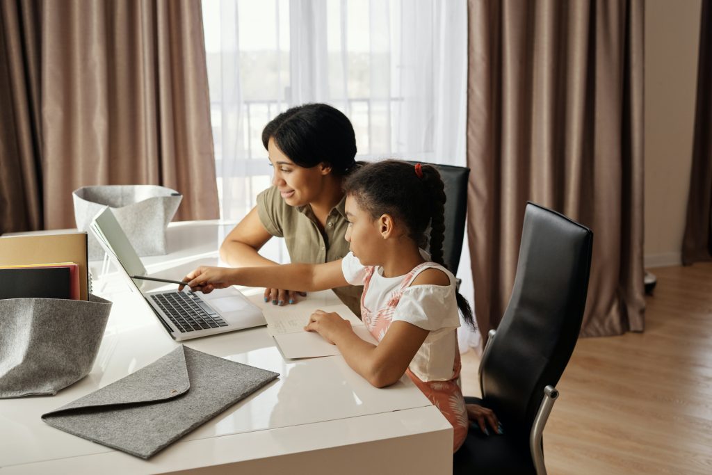Mulher ajudando uma menina a estudar no computador, demonstrando a importância da relação família e escola nas aulas online.