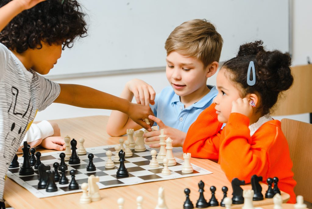 Três crianças (dois meninos e uma menina) jogando xadrez, demonstrando opções de matemática lúdica.
