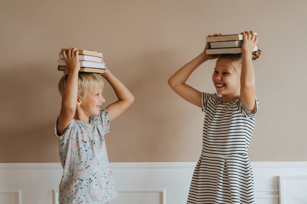 Duas crianças, um menino e uma menina, brincando de empilhar livros didáticos Ensino Fundamental sobre suas próprias cabeças. 