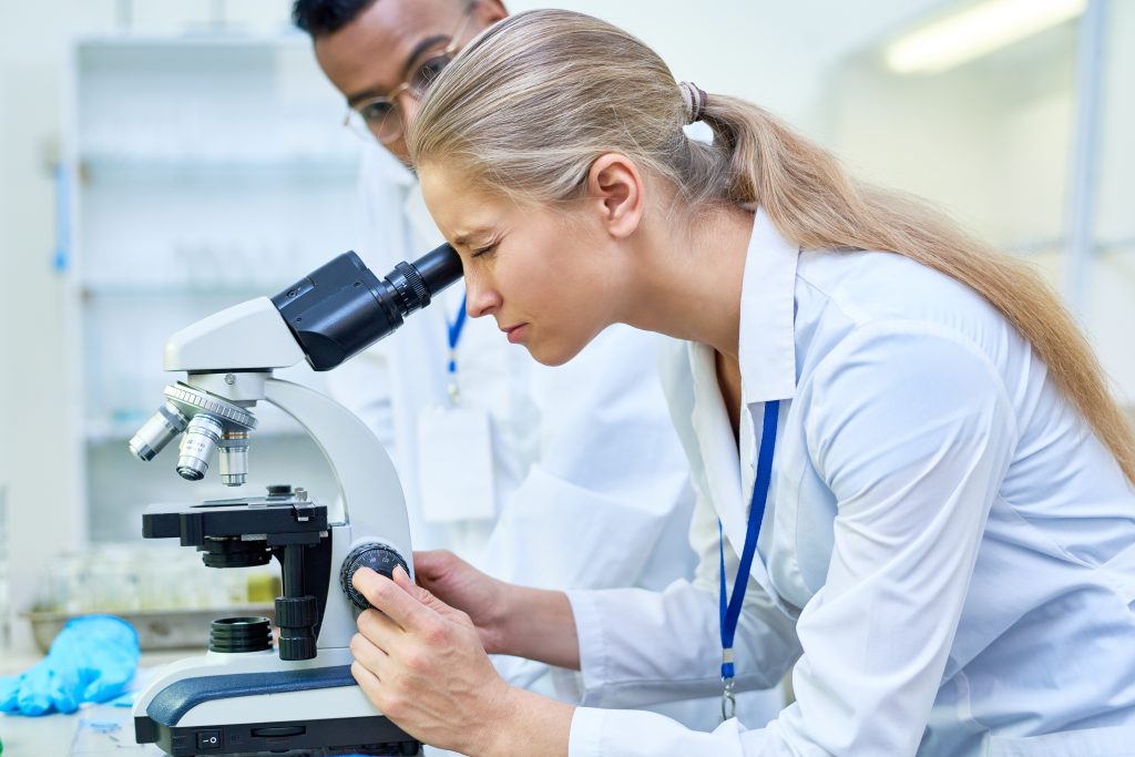 Dois estudantes no laboratório de ciência, a moça está olhando no microscópico, enquanto o rapaz a observa, em outro exemplo de disciplina que pode ser trabalhada com aprendizagem baseada em projetos. 