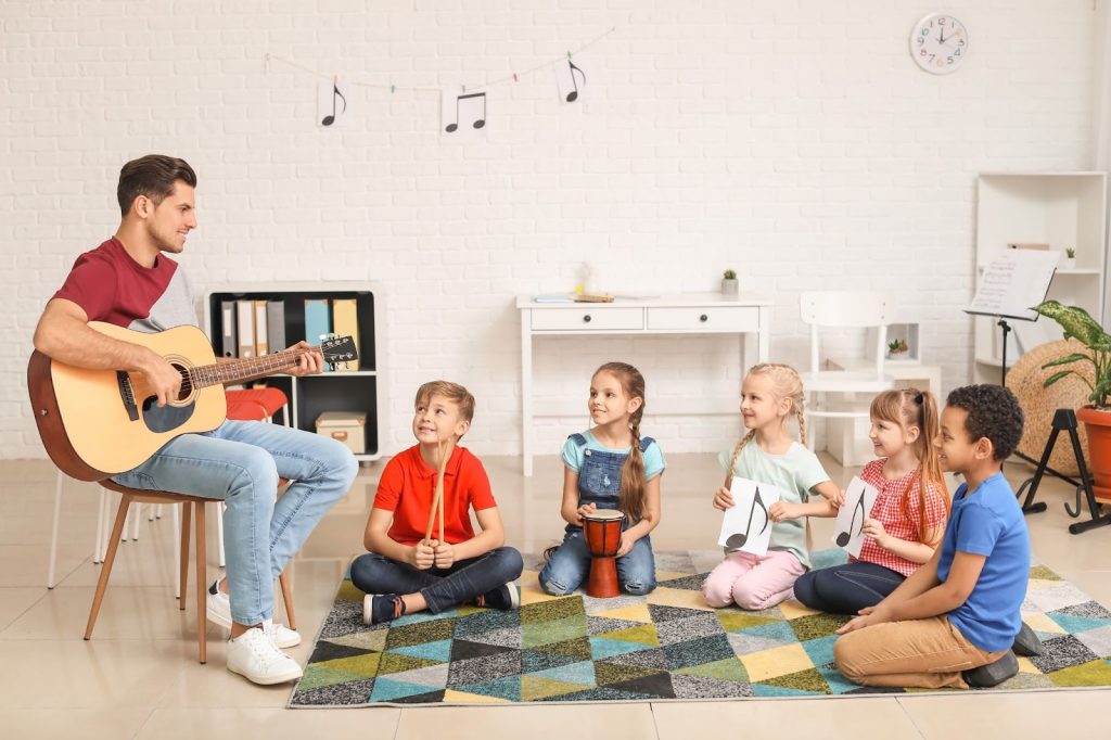 Homem sentado segurando um violão, cercado por crianças sentadas no chão, segurando instrumentos ou papéis com notas musicais para representar a musicalização na educação infantil
