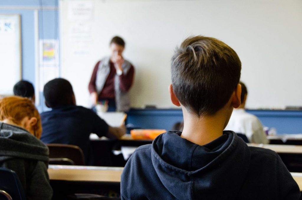 Imagem de um menino em sala de aula, ele está de costas para a foto, olhando para a frente onde está seu professor.
