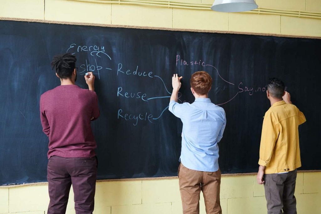 Imagem de três rapazes de costas escrevendo em uma lousa de giz. A escrita se complementa e a imagem representa um trabalho em equipe ou colaborativo